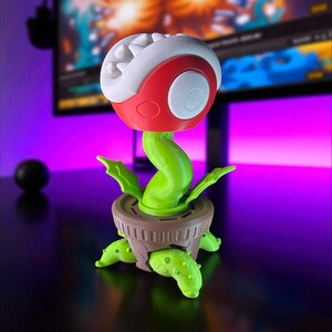May include: A novelty plant-shaped desk lamp. The lamp features a red and white head with a button, a green stem, and a brown pot base. The lamp is designed to resemble a cartoon plant.