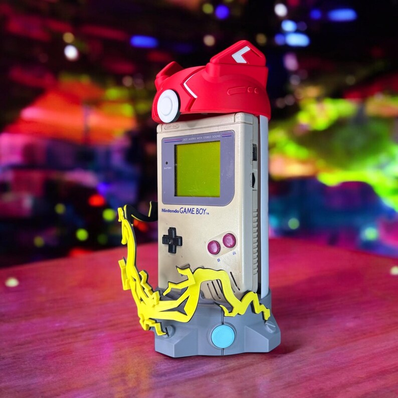 K&ouml;nnte beinhalten: Ein grauer und gelber St&auml;nder mit einem blauen Knopf h&auml;lt einen Vintage Game Boy mit einem rot-wei&szlig;en 3D-gedruckten Helm obenauf. Der Game Boy Bildschirm ist gr&uuml;n und der Text "Nintendo GAME BOY" ist sichtbar.
