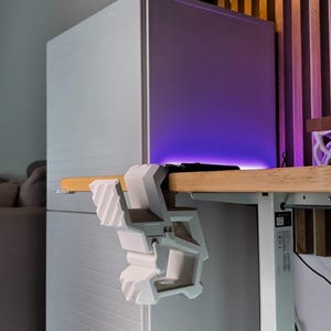 May include: A white 3D-printed desk clamp is attached to a wooden desk. The clamp is holding a small white object. The text "3D FACTORY BOX" and "o sometimes LESS is MORE &bull;" are visible in the image.