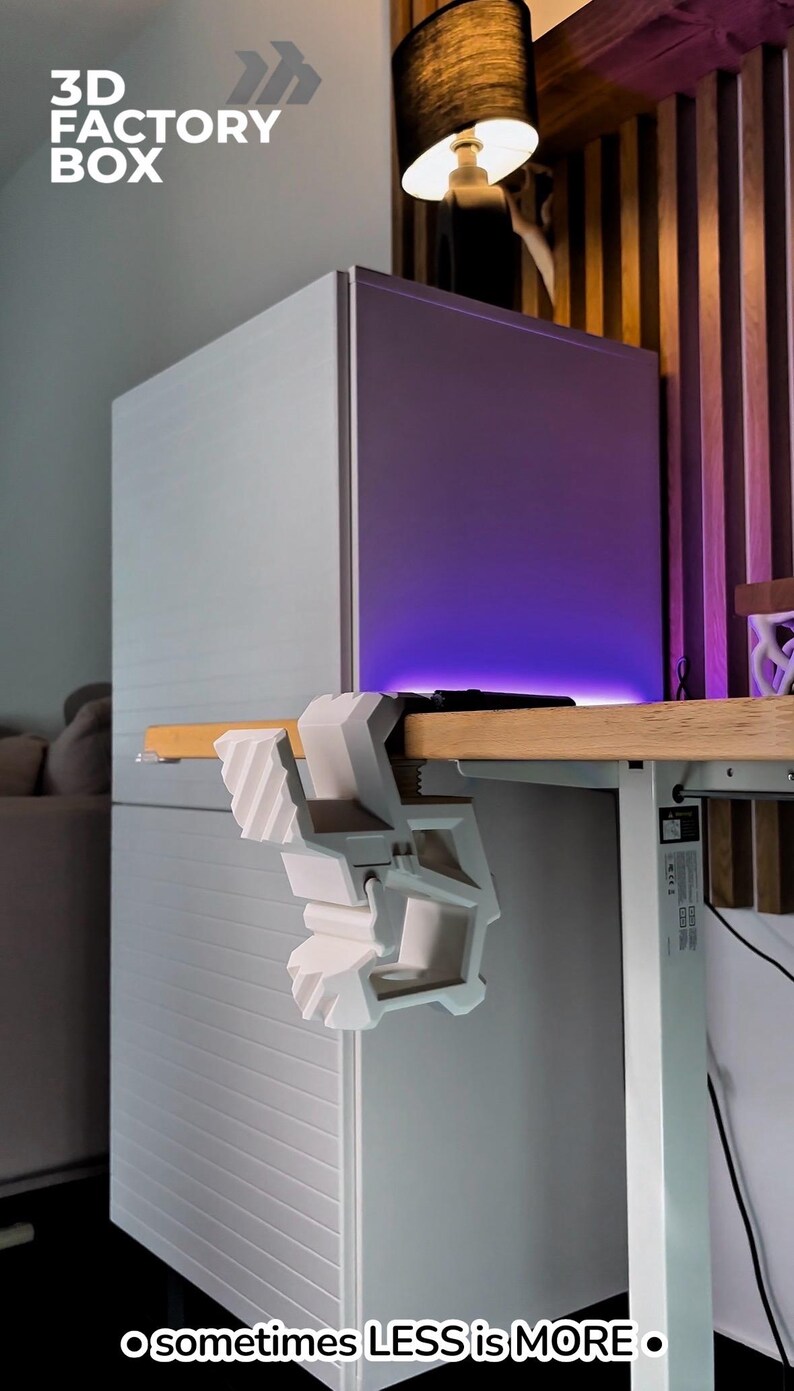 May include: A white 3D-printed desk clamp is attached to a wooden desk. The clamp is holding a small white object. The text "3D FACTORY BOX" and "o sometimes LESS is MORE &bull;" are visible in the image.