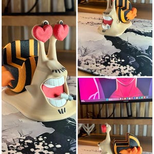 May include: A whimsical 3D-printed snail figurine with heart-shaped eyes and a wide, open mouth. The snail's shell features black and yellow stripes, and the figurine has a beige body. The base of the figurine is on a patterned surface. The text "3D FACTORY BOX" is visible.