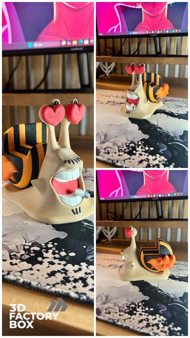 May include: A whimsical 3D-printed snail figurine with heart-shaped eyes and a wide, open mouth. The snail's shell features black and yellow stripes, and the figurine has a beige body. The base of the figurine is on a patterned surface. The text "3D FACTORY BOX" is visible.