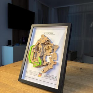 May include: A framed 3D map of a video game level, labeled "OVERPASS." The map features a detailed, multi-layered design with tan and green elements, set against a white background. The frame is black, and the map is displayed on a wooden surface.