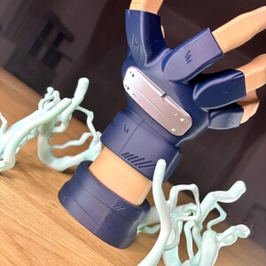 May include: A detailed figurine of a hand, featuring a blue glove with a silver plate and intricate detailing. The hand is posed with fingers extended, and surrounded by light blue, stylized energy effects. The figurine is set on a wooden surface.
