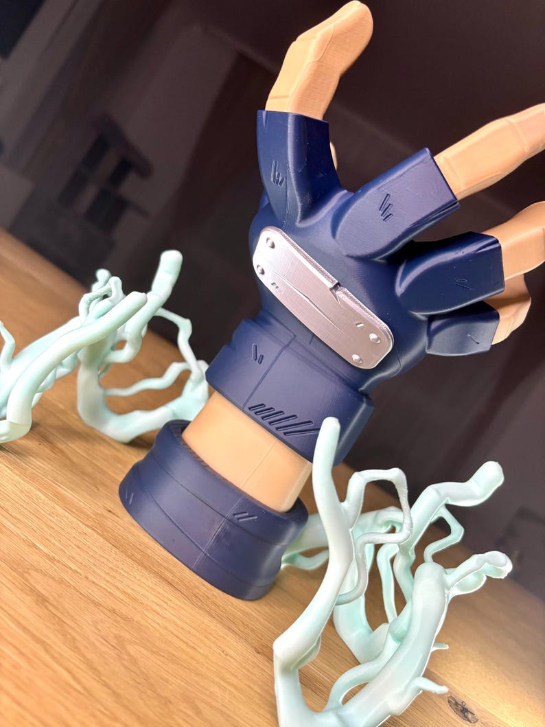 May include: A detailed figurine of a hand, featuring a blue glove with a silver plate and intricate detailing. The hand is posed with fingers extended, and surrounded by light blue, stylized energy effects. The figurine is set on a wooden surface.
