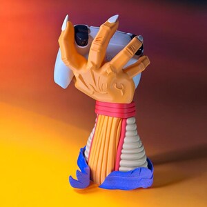 Puede incluir: Soporte de mando de juego original con forma de mano naranja que sostiene un mando blanco. Las u&ntilde;as son blancas. La base del soporte es multicolor, con rojo, amarillo y azul.