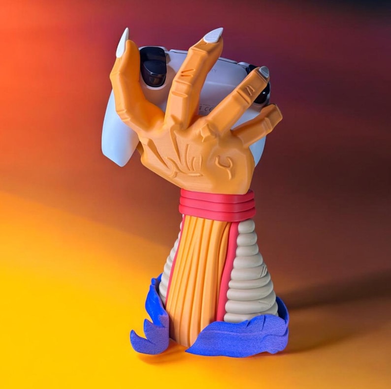 Puede incluir: Soporte de mando de juego original con forma de mano naranja que sostiene un mando blanco. Las u&ntilde;as son blancas. La base del soporte es multicolor, con rojo, amarillo y azul.