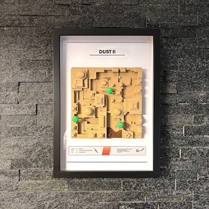 May include: A framed map of the Dust II map from the video game Counter-Strike: Global Offensive. The map is made of brown cardboard and features green trees and a red and white stripe.