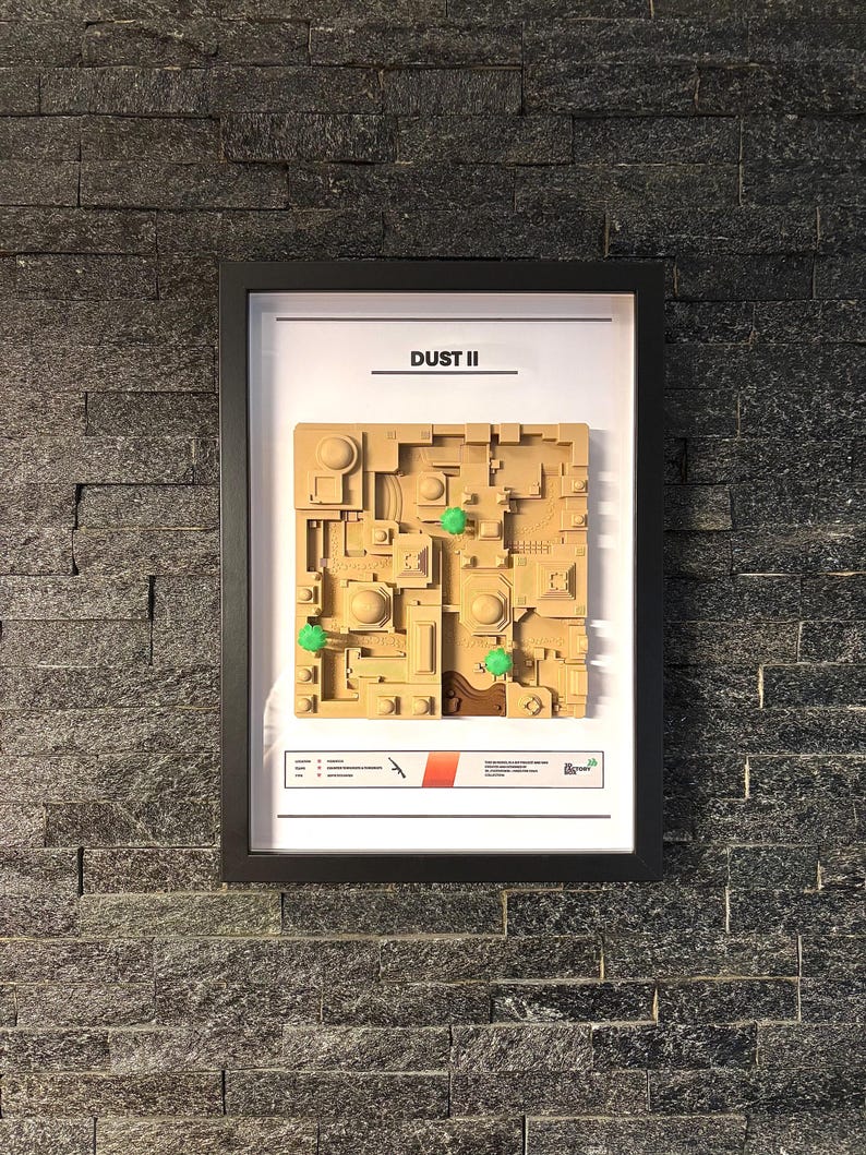 May include: A framed map of the Dust II map from the video game Counter-Strike: Global Offensive. The map is made of brown cardboard and features green trees and a red and white stripe.