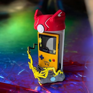 K&ouml;nnte beinhalten: Ein gelber Game Boy Color mit einer Pikachu-Spielkassette, einem roten und wei&szlig;en Pokemon-Charakterkopf und einem gelben Blitz-Design auf einem grauen Sockel.