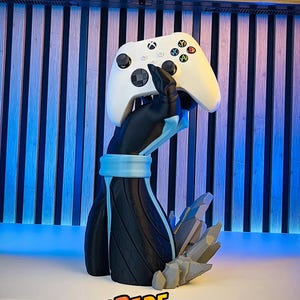 May include: A white Xbox controller rests on a black and blue 3D-printed stand. The stand features a stylized arm design with a blue band and a crystal-like base. The text "UPGRADE YOUR SETUP!" is displayed.