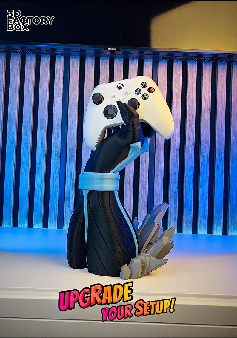 May include: A white Xbox controller rests on a black and blue 3D-printed stand. The stand features a stylized arm design with a blue band and a crystal-like base. The text "UPGRADE YOUR SETUP!" is displayed.