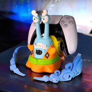 May include: A blue and orange cartoon slug holding a white PlayStation 5 controller. The slug is wearing a green and orange outfit with a black and white design. The controller is resting on the slug's back. There are two blue plastic pieces on the table.