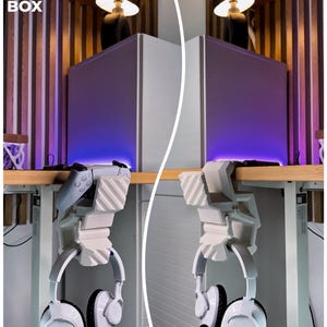 May include: White gaming headset and controller hanging from a white 3D-printed desk mount. The mount is attached to a wooden desk. The text "3D FACTORY BOX" is in the upper left corner.