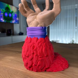 May include: A 3D printed figurine of a hand with a red, textured base and a purple band around the wrist.