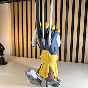 May include: A 3D printed figurine of a blue and yellow gloved hand with three long silver claws extending upward. The hand is mounted on a jagged gray base. The text 3D FACTORY BOX is visible in the bottom right corner of the image.