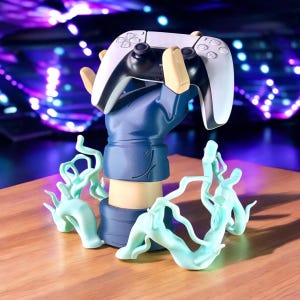 Könnte beinhalten: Ein Gaming-Controller, der von einem stilisierten, handförmigen Ständer gehalten wird. Der Ständer ist blau und beige, mit hellblauen dekorativen Akzenten. Der Controller ist schwarz und weiß. Der Hintergrund hat einen verschwommenen lila und blauen Lichteffekt.