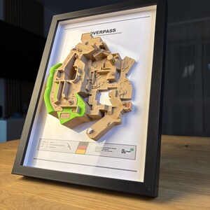 May include: Framed 3D map of the Overpass game level, featuring tan and green terrain details. The map is set against a white background within a black frame. The frame is angled on a wooden surface.
