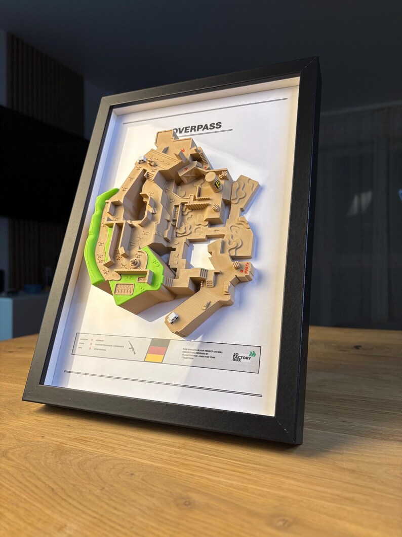 May include: Framed 3D map of the Overpass game level, featuring tan and green terrain details. The map is set against a white background within a black frame. The frame is angled on a wooden surface.