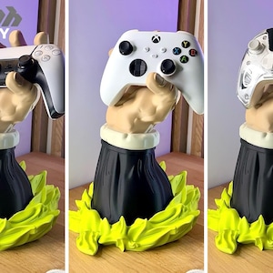 Op de afbeelding: Een 3D-geprinte gamecontrollerhouder met een hand die een wit-zwarte PlayStation 5-controller, een witte Xbox-controller en een zilveren controller vasthoudt. De basis is zwart met een witte kraag en een groen bladontwerp.