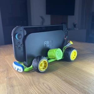 K&ouml;nnte beinhalten: Eine schwarze Nintendo Switch-Konsole ist auf einem gr&uuml;n-gelben 3D-gedruckten Spielzeugauto-Chassis montiert. Das Auto hat schwarze R&auml;der mit gelben Zentren und einen gr&uuml;nen Tankdeckel. Auf dem Bildschirm der Konsole ist das Nintendo-Logo zu sehen.