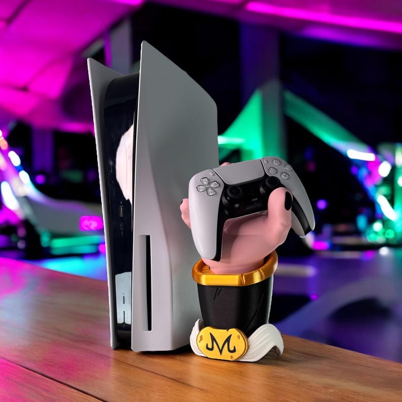 May include: A white and black PlayStation 5 console stands next to a controller held by a hand-shaped stand. The stand is black with gold accents and a white base with a gold emblem. The controller is gray and black.
