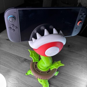 May include: A green, red, and white 3D-printed phone or tablet stand shaped like a Piranha Plant from the Super Mario video game series. The stand is holding a black Nintendo Switch console. The stand has a brown base and green leaves.