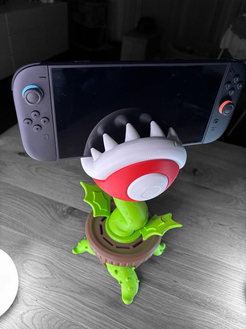 May include: A green, red, and white 3D-printed phone or tablet stand shaped like a Piranha Plant from the Super Mario video game series. The stand is holding a black Nintendo Switch console. The stand has a brown base and green leaves.