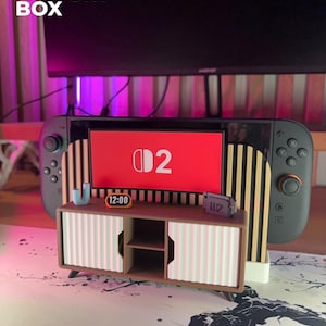 May include: A 3D-printed miniature entertainment center with a Nintendo Switch console. The center features a brown cabinet with pink and white striped doors, a small clock, and a miniature game cartridge. The Switch screen displays the number "2".