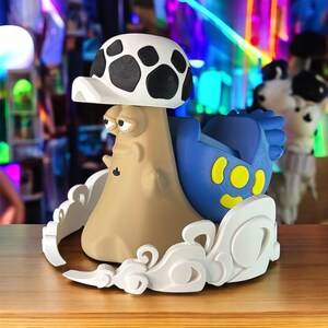 May include: A whimsical mushroom-shaped figurine with a tan body, a white and black spotted cap, and a blue character with yellow accents. The figurine is set on a white cloud-like base, creating a playful and unique decorative piece.