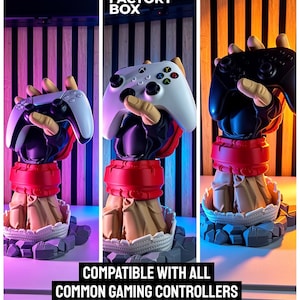 Puede incluir: Un soporte con forma de mano esculpida con una mu&ntilde;equera roja y una base de tela color canela, dise&ntilde;ado para sostener mandos de juego. El soporte se muestra con mandos blancos y negros. El texto dice 3D FACTORY BOX y COMPATIBLE WITH ALL COMMON GAMING CONTROLLERS.