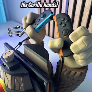 May include: A gaming accessory featuring a gorilla hand design holding two Joy-Con controllers. The text reads, "Put your 3rd and 4th JoyCon into the Gorilla hands!" The controllers are black and blue.