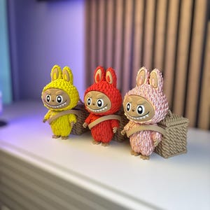 May include: Three colorful figurines in yellow, red, and pink, each with a bunny-like hood and a smiling face. They are standing in front of a woven basket. The figurines have a unique, cartoonish design.