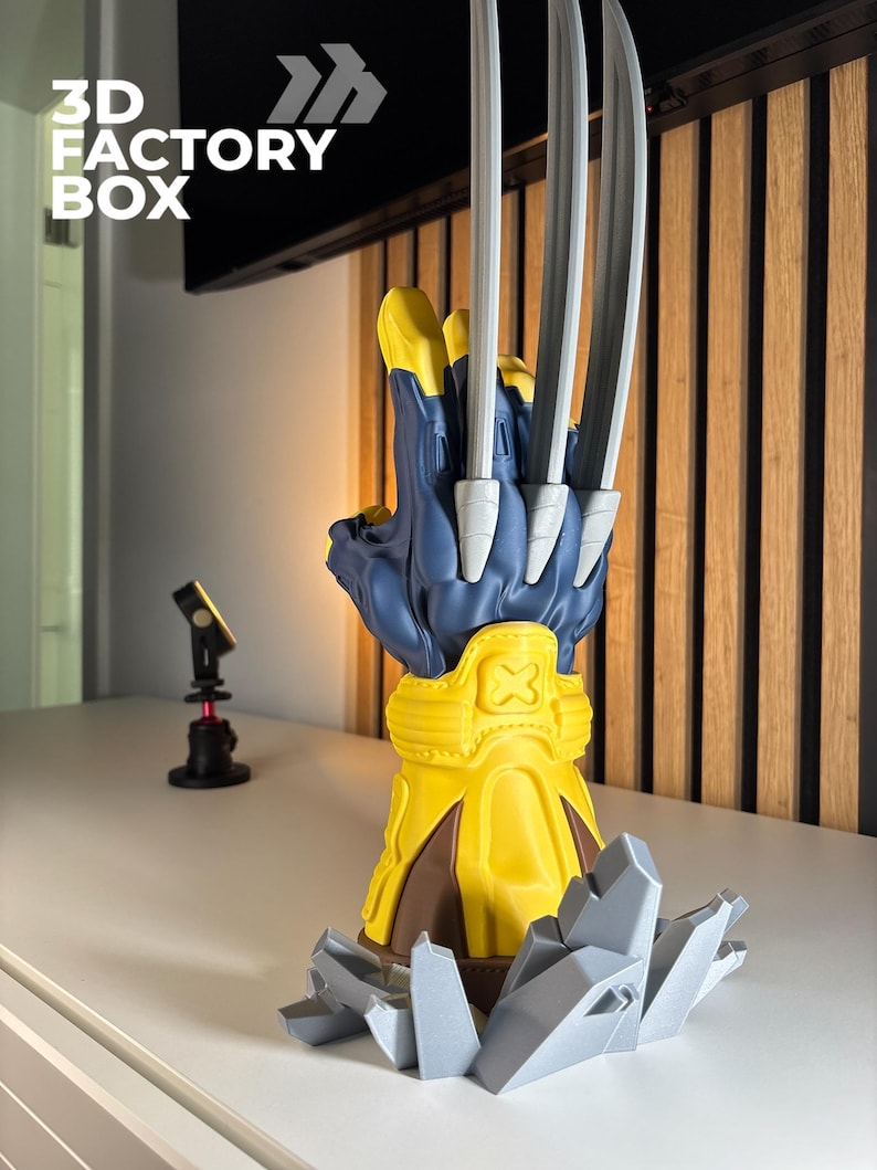May include: A 3D printed display model of a blue and yellow glove with three long silver claws extending from the knuckles. The glove features a yellow wrist cuff with an X emblem and rests on a base of jagged, light gray rock-like structures.