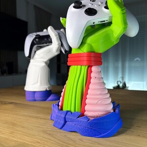 Puede incluir: Un soporte para mando de videojuegos multicolor con forma de mano, que sostiene un mando blanco. El soporte es verde, rojo, rosa y azul. En el fondo, hay otro soporte que sostiene un mando en blanco y negro.