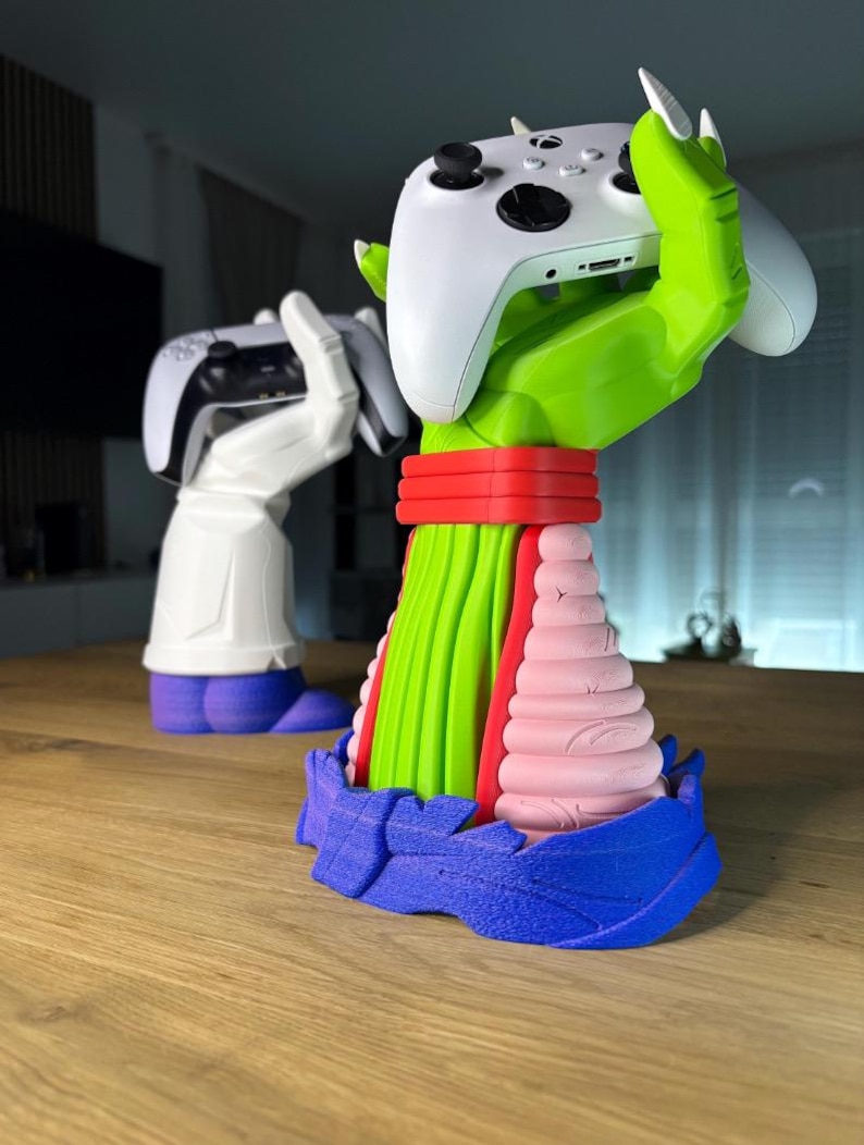 Puede incluir: Un soporte para mando de videojuegos multicolor con forma de mano, que sostiene un mando blanco. El soporte es verde, rojo, rosa y azul. En el fondo, hay otro soporte que sostiene un mando en blanco y negro.
