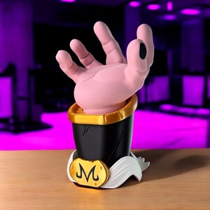 May include: A pink hand figure with a black base and gold trim. The figure has a white and gold emblem with the letter "M" on the front. The hand is in a gesture, with the fingers slightly curved.