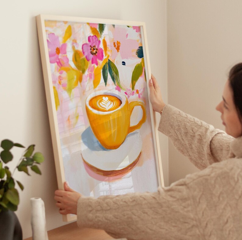 May include: Framed art print featuring a vibrant yellow coffee mug with latte art, set against a floral background of pink, yellow, and green. The artwork is displayed in a light-colored wooden frame, perfect for kitchen or dining room decor.