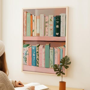 May include: A framed print of a bookshelf filled with colorful books. The books are in shades of pink, blue, green, and cream, with floral and botanical designs. The bookshelf is painted pink and the frame is a light wood color.