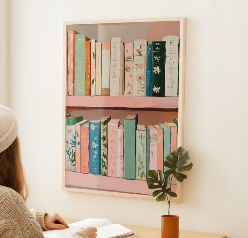 May include: A framed print of a bookshelf filled with colorful books. The books are in shades of pink, blue, green, and cream, with floral and botanical designs. The bookshelf is painted pink and the frame is a light wood color.