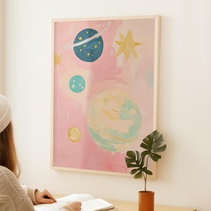 May include: A pink and blue abstract painting of planets and stars. The painting is framed in a light wood frame.