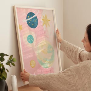 May include: A framed abstract painting with a pink background and colorful planets and stars. The painting is in a white frame and is being hung on a wall.