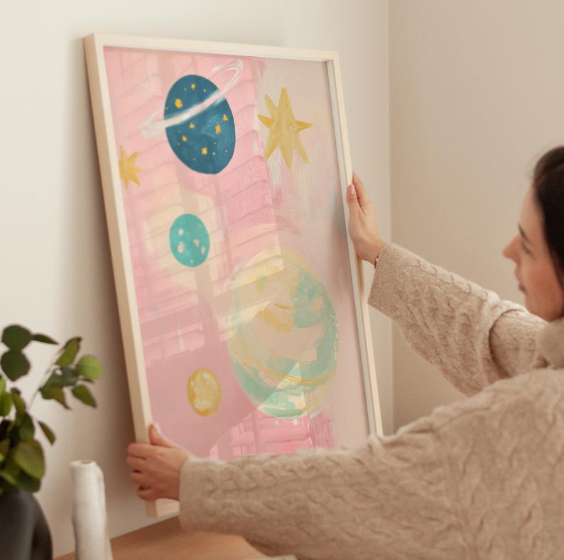 May include: A framed abstract painting with a pink background and colorful planets and stars. The painting is in a white frame and is being hung on a wall.