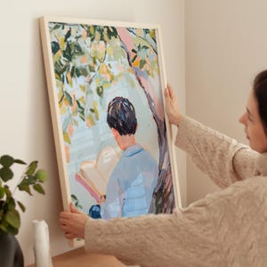 Puede incluir: Una pintura enmarcada de una persona sentada bajo un &aacute;rbol y leyendo un libro. La pintura est&aacute; en un marco de madera claro y tiene un esquema de color azul claro, verde y blanco.