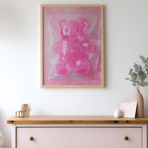 May include: A framed pink gummy bear sculpture in a clear plastic bag. The bear is sitting on a pink background. The frame is made of wood and has a light brown finish.