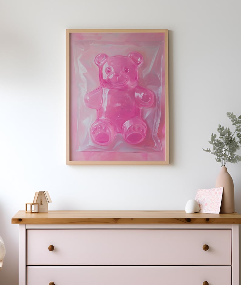 May include: A framed pink gummy bear sculpture in a clear plastic bag. The bear is sitting on a pink background. The frame is made of wood and has a light brown finish.