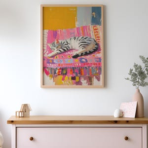 May include: A framed painting of a gray and white cat sleeping on a colorful rug. The rug has pink, yellow, blue, and green stripes and patterns. The background is a white wall.