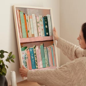 May include: A framed print of a bookshelf filled with colorful books. The books have various colors and patterns, including floral and solid designs. The frame is white, and the print is being held by a person.