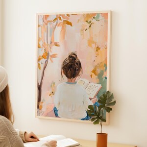May include: A framed art print showing a person with a bun reading a book, seen from the back. The artwork uses a soft palette of pinks, oranges, and greens, with a tree and foliage in the background. The frame is a light wood colour.
