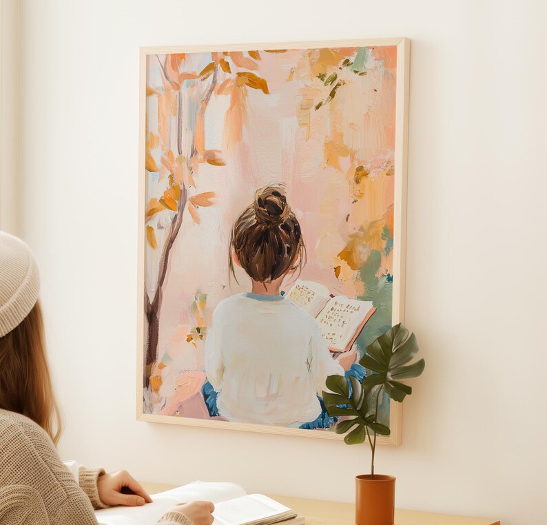 May include: A framed art print showing a person with a bun reading a book, seen from the back. The artwork uses a soft palette of pinks, oranges, and greens, with a tree and foliage in the background. The frame is a light wood colour.